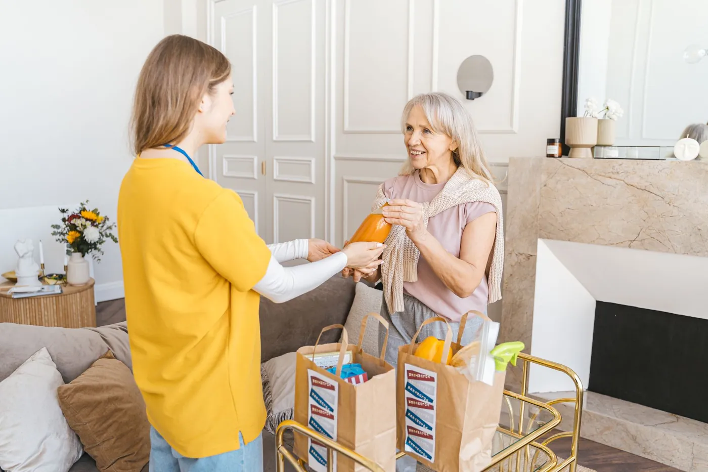 A caregiver hands a smiling older woman a bottle from grocery bags brought into her living room