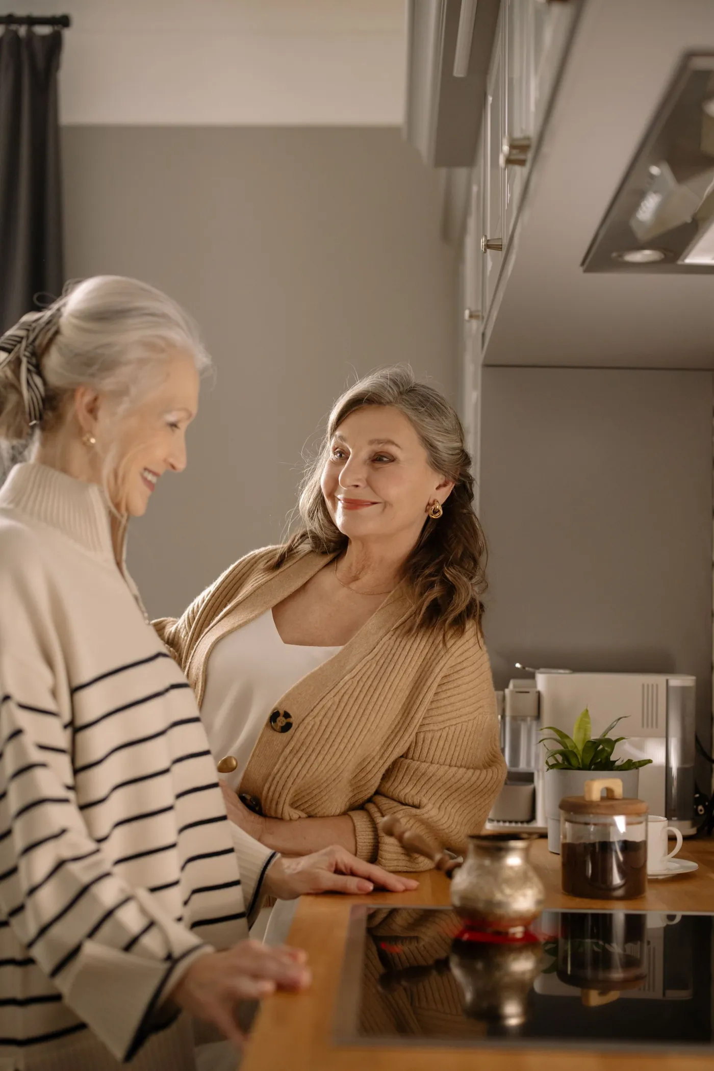 Two older women smiling at each other over coffee in a bright home kitchen