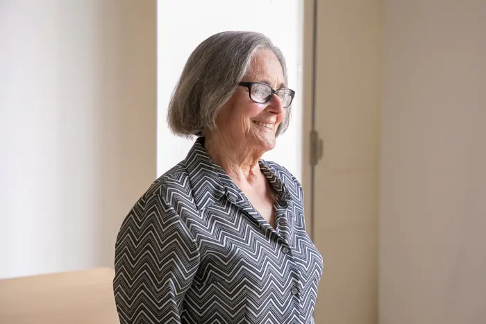 A smiling older woman in glasses looking out of a sunny window at home